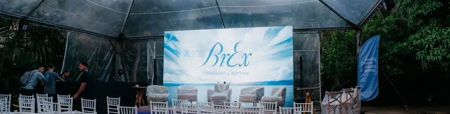 Telão exibindo o logotipo cursivo "BrEx" em área de convivência ao ar livre